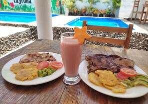 Comida con carne a la parrilla, plátanos y aguacate sobre una mesa de madera, con una piscina y sillas al fondo.