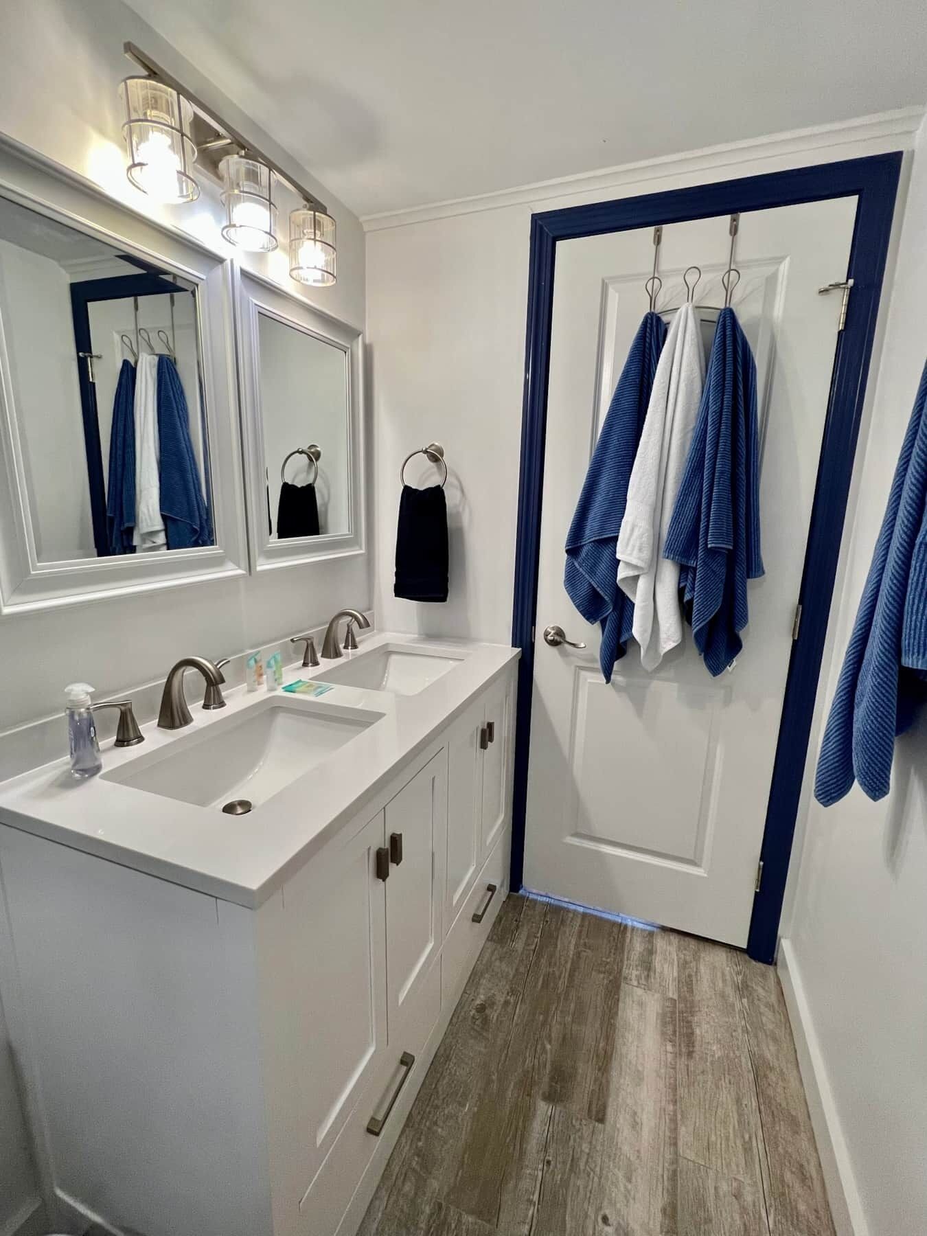 Un baño con dos lavamanos, un espejo y una puerta con toallas colgadas en ganchos.