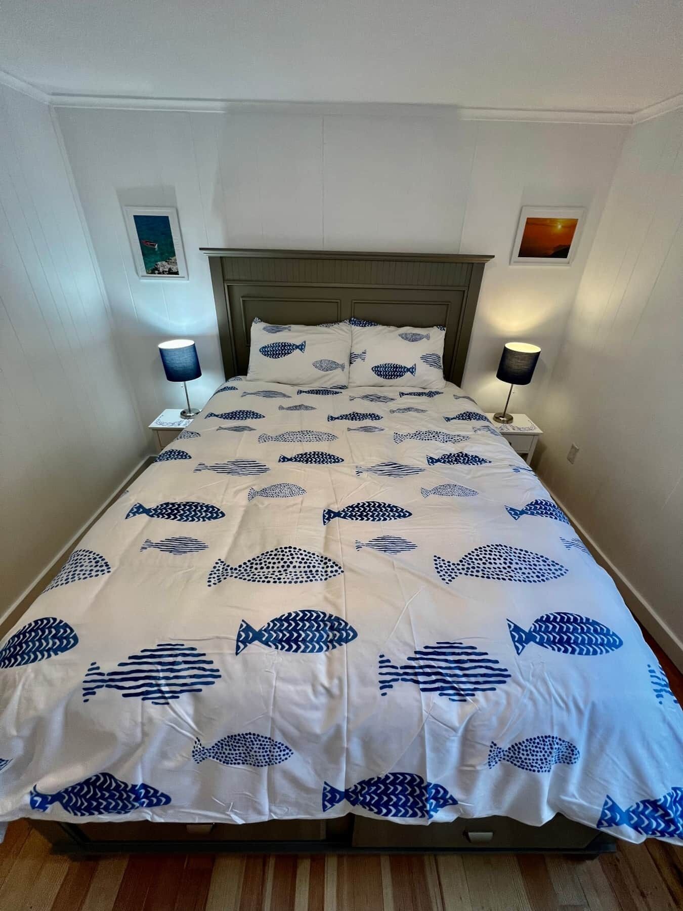 A bed with a white bedspread featuring blue fish patterns, flanked by two bedside lamps and framed pictures on the walls.