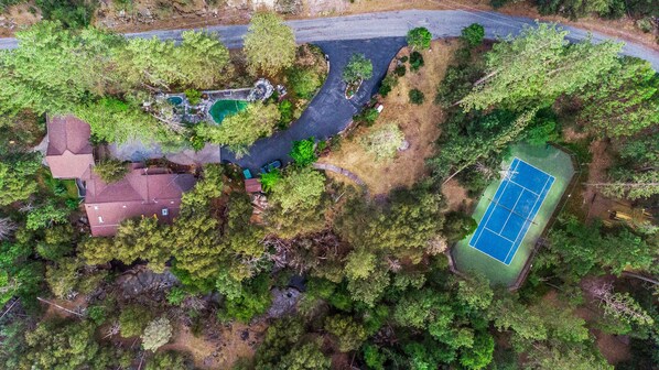 An aerial view of a residential area with a tennis court, a swimming pool, and a driveway leading to a house with a red roof. - Riverfront Paradise w/ Pool, Spa & Tennis Court (Oakhurst)