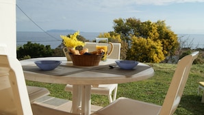 A round table with a basket of croissants, a glass of orange juice, and two bowls. The table is outdoors with a view of the sea and trees in the background. - Les Cyprès - View to the sea and Harbor of Santa Severa, in Corsican Cape (Luri)