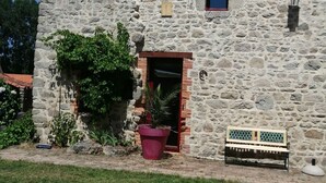 A stone building with a red-tiled roof, a small window, and a door. There is a bench and a potted plant in front.