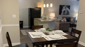 A dining area with a dark wood table set for four, surrounded by matching chairs. A kitchen with a refrigerator and sink is visible in the background. - Fabulous One Bed/One Bath Corporate Furnished Apt in Alpharetta (Alpharetta)