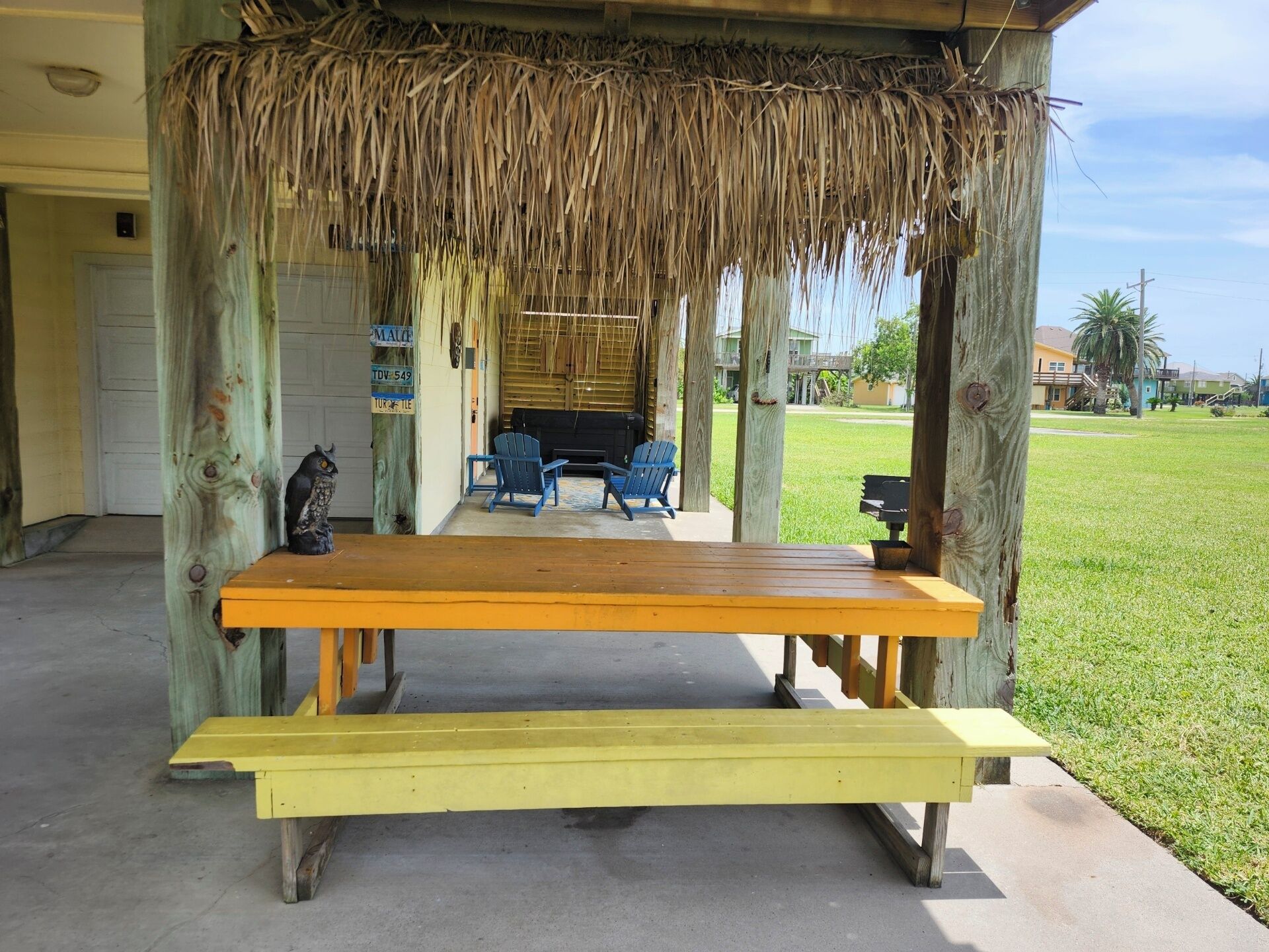 A covered outdoor seating area with a bench and chairs, surrounded by wooden posts and a thatched roof.