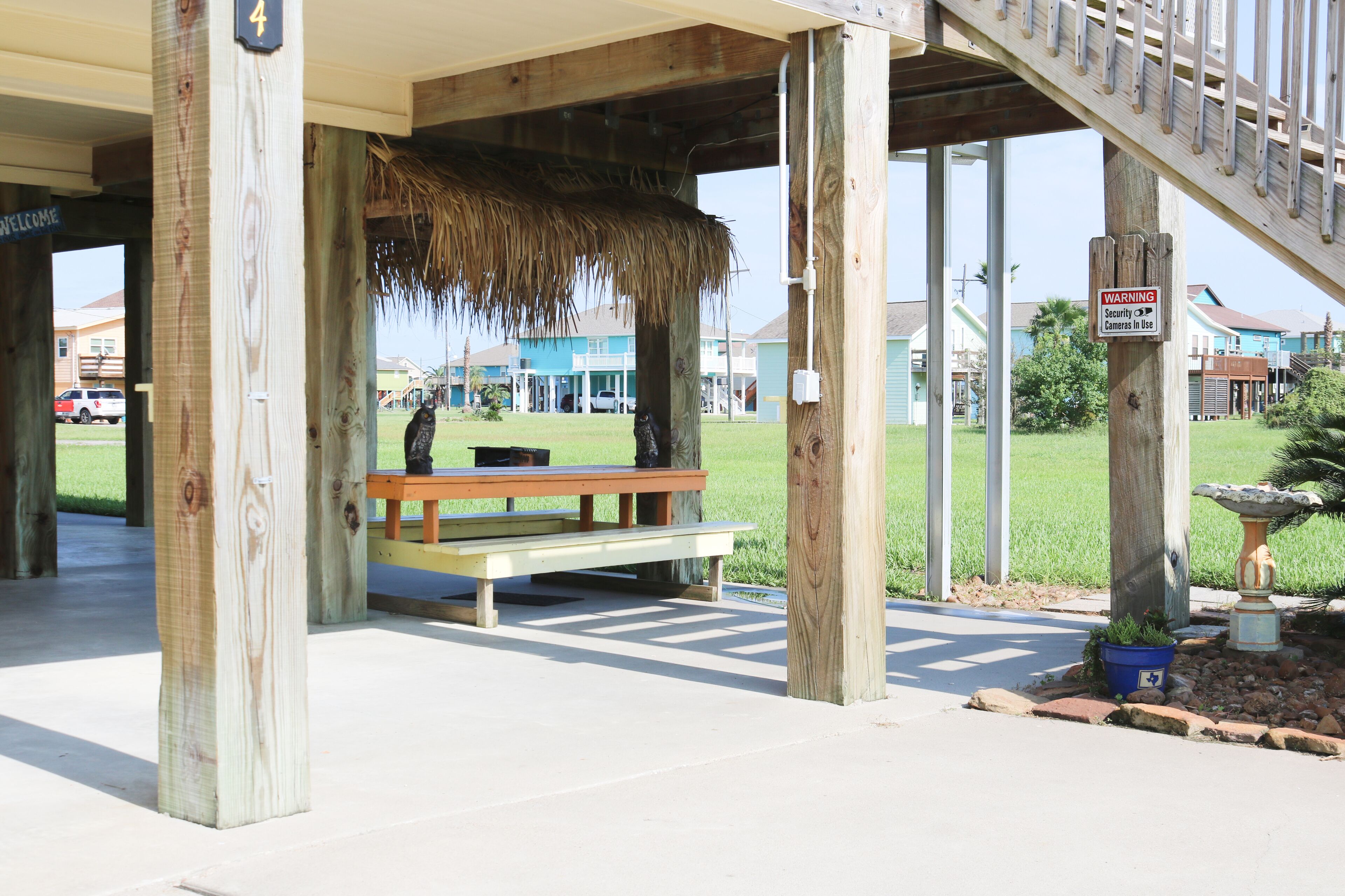 A covered outdoor area with a wooden bench, a thatched roof, and a warning sign about security cameras.