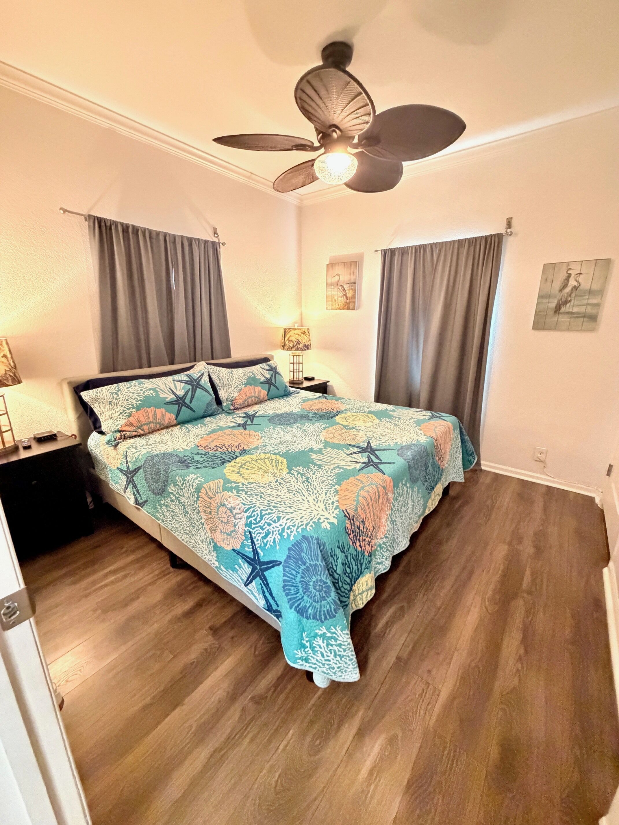 Une chambre à coucher comprenant un lit, un ventilateur de plafond, une table de chevet, une lampe et deux tableaux accrochés au mur.
