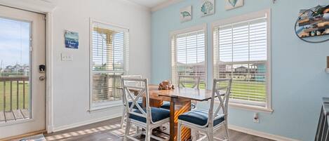 A dining area with a table and chairs, large windows with blinds, and a view of a green landscape.