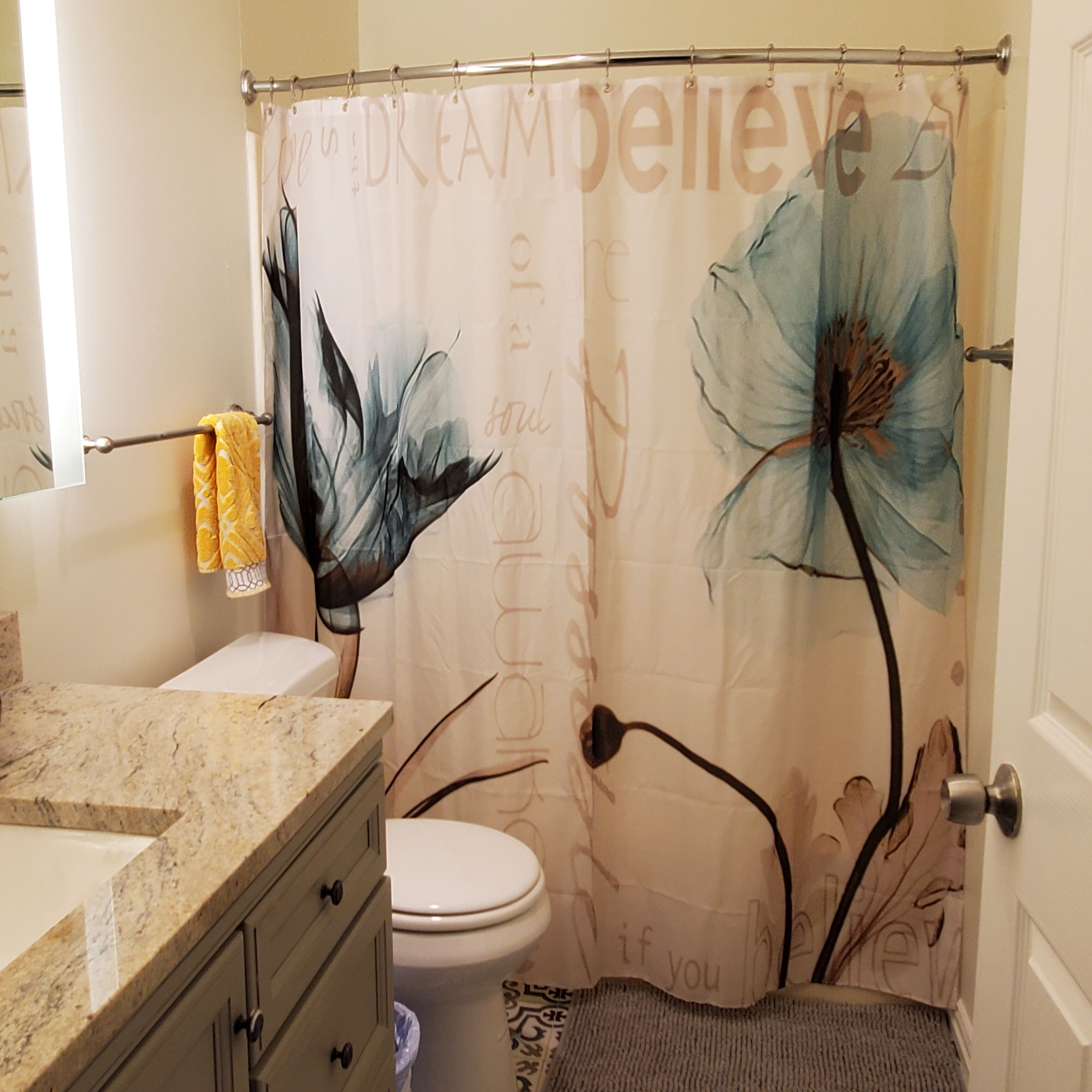 A bathroom with a shower curtain featuring floral designs, a toilet, a sink with a marble countertop, and a towel rack with yellow towels.