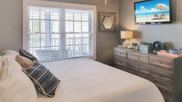 A bedroom with a large bed, a dresser, a TV on the wall, and a window with blinds. - Beach house with pool only steps from the ocean (Murrells Inlet)