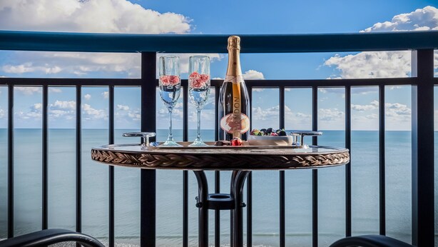 A table with a bottle of champagne, two glasses, and a bowl of fruit on a balcony with a view of the sea.