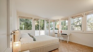 Ein Schlafzimmer mit Bett, Schreibtisch und Stuhl, großen Fenstern und Blick auf Bäume.