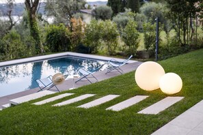 Un espace piscine offrant une vue dégagée sur l’eau, une chaise longue et deux lampes sphériques illuminées posées sur la pelouse.