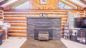 A log cabin interior with a stone fireplace, wooden beams, a television, and a bookshelf. - Rustic Roost Log Cabin | 1 Bed, 1 Bath (Custer)