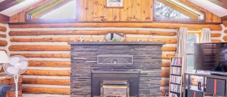 A log cabin interior with a stone fireplace, wooden beams, a television, and a bookshelf.