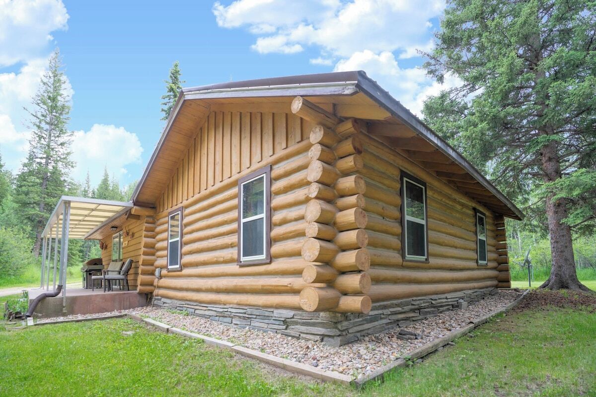A log cabin with a porch, a stone foundation, and a view of the surrounding forest.