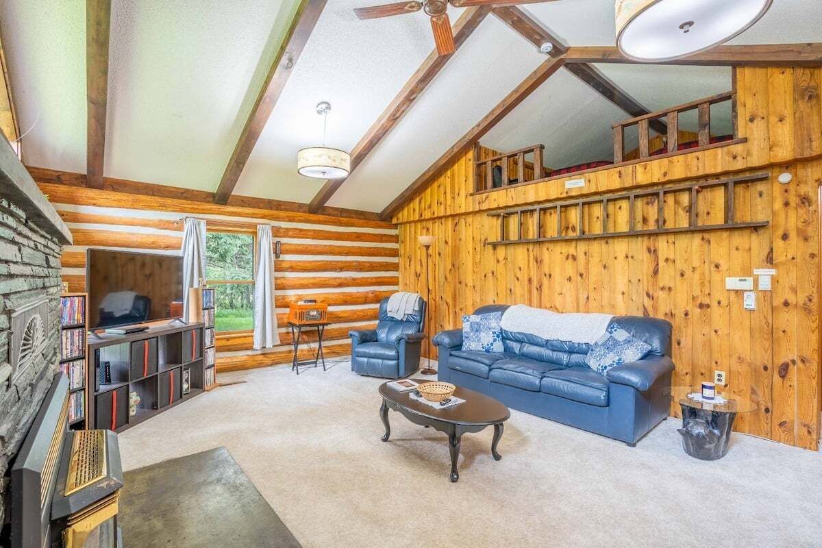 A log cabin interior with a sofa, armchairs, a coffee table, a fireplace, and a bookshelf.