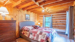 A log cabin bedroom with a bed, dresser, and windows. - Rustic Roost Log Cabin | 1 Bed, 1 Bath (Custer)