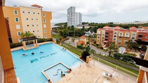 A swimming pool with people, surrounded by apartment buildings and a view of a tall building in the distance.