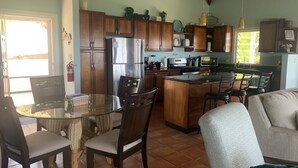 A kitchen with wooden cabinets, a stainless steel refrigerator, a microwave, and a dining area with a glass table and chairs.