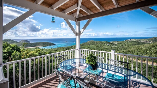 A covered outdoor balcony with a glass table and chairs, offering a view of a coastal landscape with greenery and the sea.