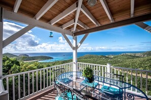 Un balcon extérieur abrité, doté d’une table et de chaises en verre, offrant une vue sur un paysage côtier verdoyant et la mer.