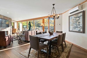 A spacious dining area with a large wooden table and chairs, a stone fireplace, and a view of autumn foliage through large windows.