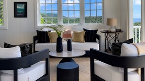 Ein modernes Wohnzimmer mit Deckenventilator, großen Fenstern mit Blick auf Grünflächen, einer Sofagarnitur und einem Couchtisch.