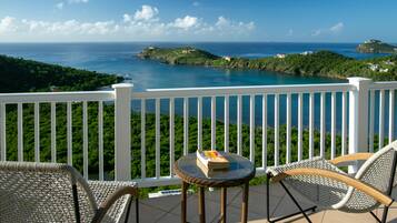 A balcony with wicker chairs and a small table, overlooking a coastal landscape with clear skies.