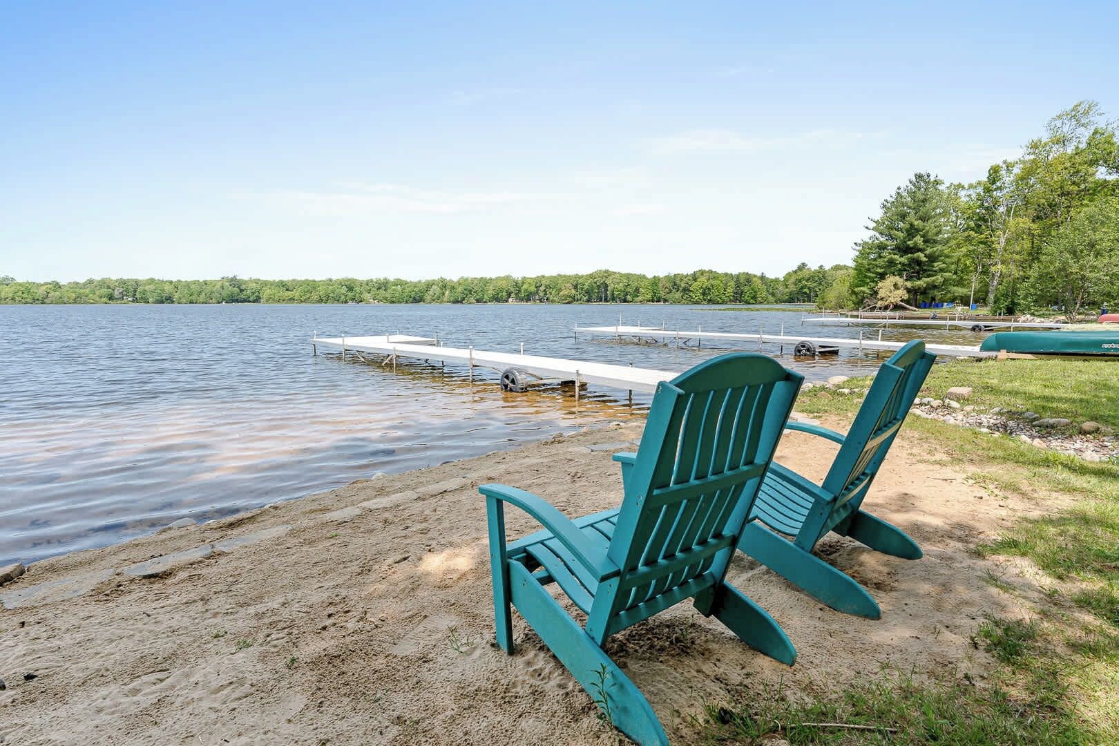 Dos sillas Adirondack verdes sobre una playa arenosa junto a un lago tranquilo con un muelle que se adentra en el agua.
