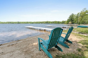 Kaksi vihreää Adirondack-tyylistä tuolia hiekkarannalla, rauhallisen järven äärellä, ja laiturin pää ulottuu veteen.