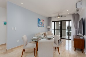 Una sala de estar moderna con una mesa de comedor de vidrio, asientos cómodos, un ventilador de techo y un ventanal con cortinas.