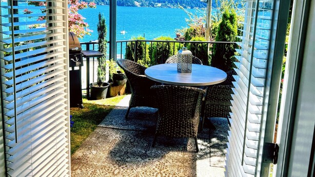 A covered outdoor seating area with a table and chairs, overlooking a lake with a forested shoreline. - Bill's Landing Luxury Suite with private Hot Tub (Gibsons)