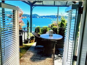 Ein überdachter Außenbereich mit Tisch und Stühlen mit Blick auf einen See und einen bewaldeten Uferstreifen.