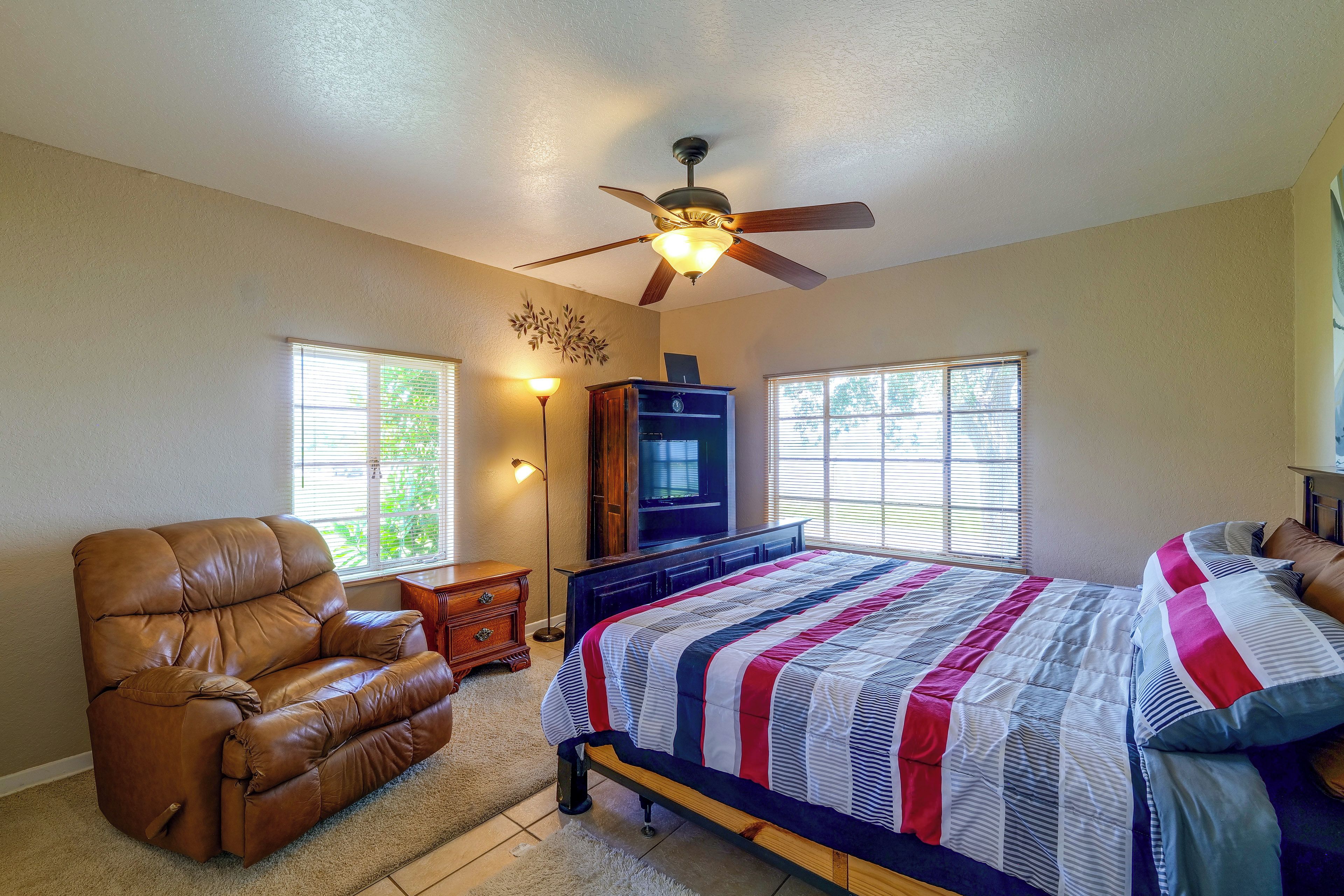 A bedroom with a bed, a leather chair, a ceiling fan, and two windows.