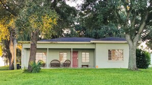 Una casa a un piano con facciata verde chiaro, tetto scuro e un portico con due sedie di legno.