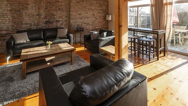 A spacious living room with a brick wall, wooden beams, a black leather sofa, a wooden coffee table, and a dining area with a table and chairs.