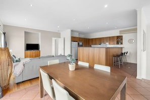 A modern kitchen with wooden cabinets, a built-in oven, and a dining area with a wooden table and white chairs. - Cowrie House: Ocean Views, 500m to Fisho’s Beach (Torquay)