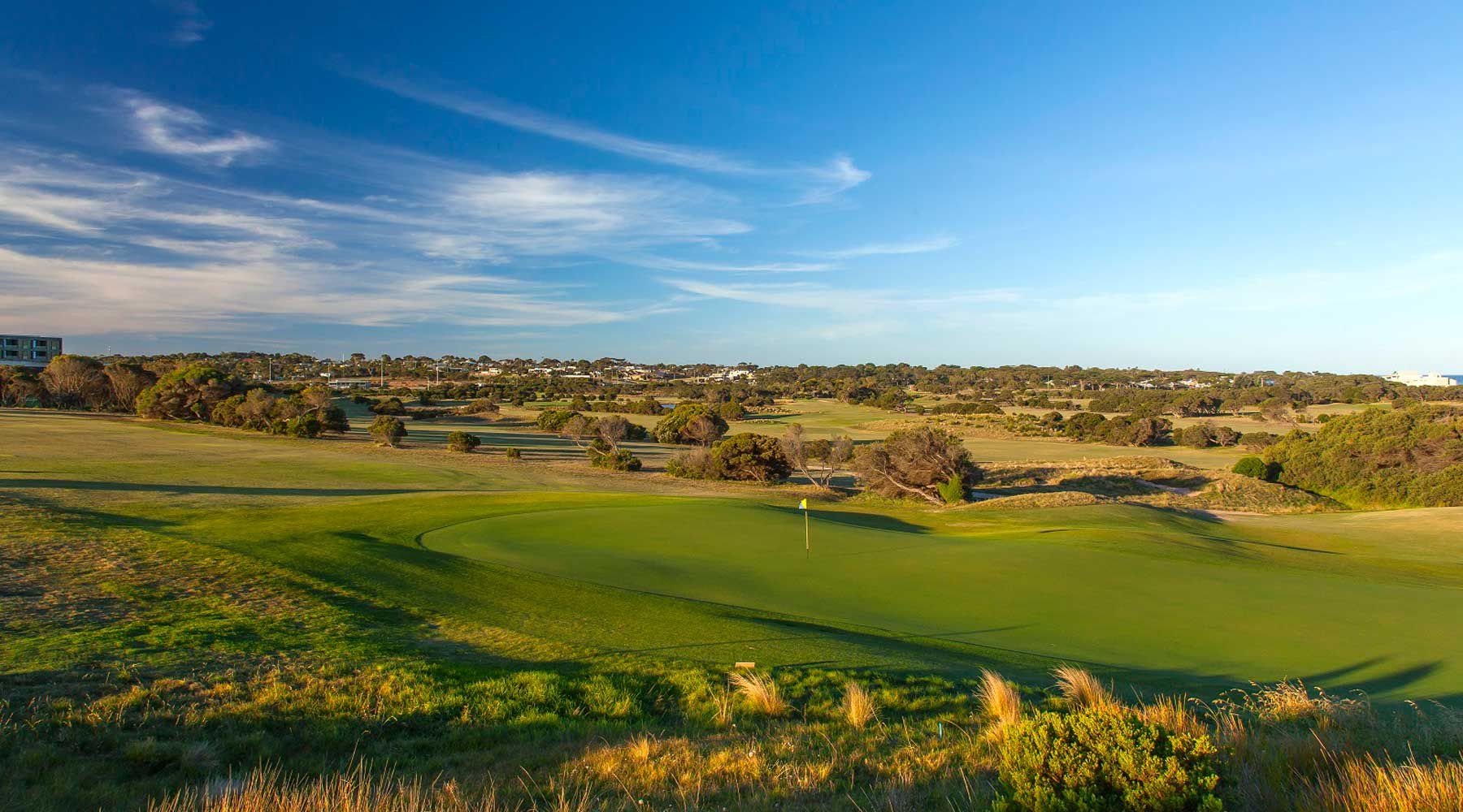 A golf course with a flag, trees, and distant buildings.