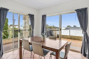 A dining area with a table and chairs, large windows with curtains, and a view of a building with solar panels. - Cowrie House: Ocean Views, 500m to Fisho’s Beach (Torquay)