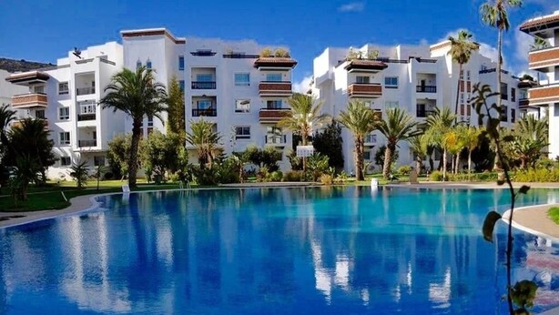 A swimming pool surrounded by multi-story buildings with balconies, palm trees, and a clear blue sky.