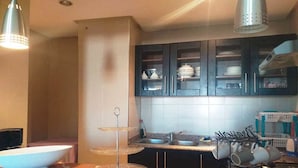 A kitchen with a white sink, a double-tiered glass serving stand, dark cabinets with glass doors, and a metal rack holding cups and plates.