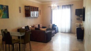A spacious living area with a dining table and chairs, a sofa set, a chandelier, and a television mounted on the wall.