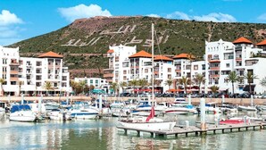 A marina with boats docked, surrounded by multi-story buildings with balconies, and a hill in the background.
