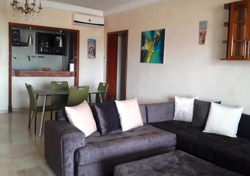 Ein Wohnzimmer mit grauem Sofa, einem Esstisch mit Stühlen und einer Küchenzeile im Hintergrund.