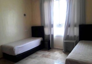 A small, single-room hotel room with two beds, a window with curtains, and a tiled floor. - Marina Agadir Residence 5419 (Agadir)