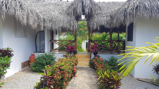 A thatched-roof structure with a central staircase leading to a covered area, surrounded by various plants and a gravel pathway. - Charmant Bungalow Tropical (Las Terrenas)