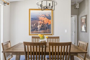A dining area with a wooden table and chairs, a framed landscape painting, and a vase with flowers.