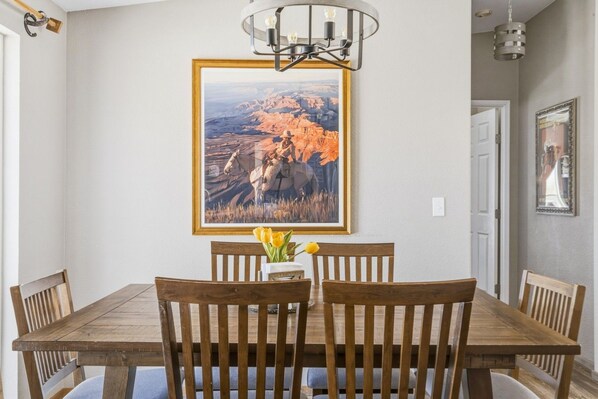 A dining area with a wooden table and chairs, a framed landscape painting, and a vase with flowers.