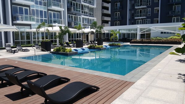 A modern hotel pool area with lounge chairs, a swimming pool, and a glass-enclosed building in the background.