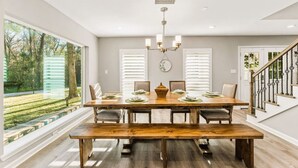 A dining room with a large wooden table set for six, flanked by large windows with white blinds, a chandelier, and a staircase with a decorative railing. - Breakfast with a View with BBQ Firepit Golf Course (Conroe)